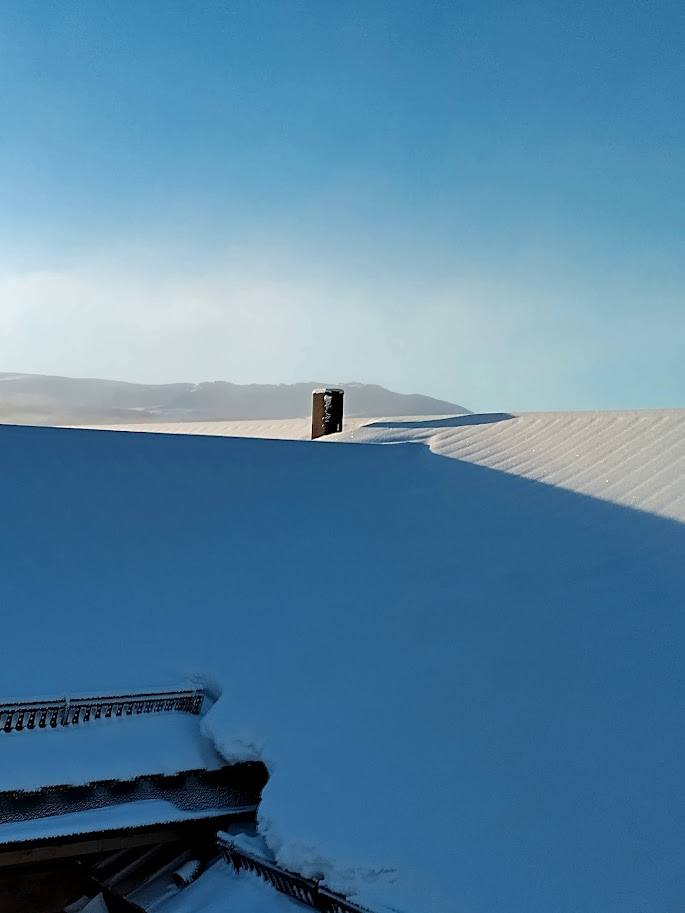 Snötäckt plåttak med skorsten mot blå himmel i Jämtland